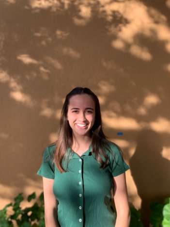 A woman with medium-length brown hair wearing a green blouse with white buttons smiles at the camera in front of a brown wall.